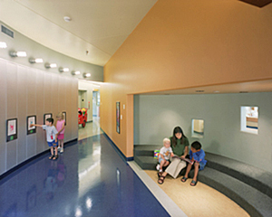 Colorful hallway with 2 children looking at pictures on the wall and a teacher and child reading in the niche built into the hall